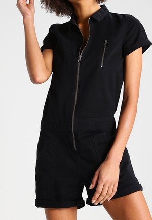 Combinaison noire à manches courtes avec fermeture éclair à l'avant, col et poche poitrine. Confectionnée en tissu uni avec poignets retroussés et poches latérales.