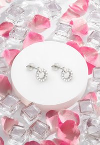 Silver cirkulära örhängen prydda med klara kristaller, visade på en vit pedestal omgiven av rosa rosenblad och isbitar.
