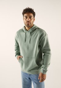 Felpa con cappuccio in misto cotone verde spento, dotata di tasca frontale, polsini a costine e lacci. Indossata con jeans azzurri chiari.