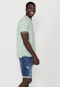 Polo di colore verde chiaro con dettagli bianchi, maniche corte e piccoli motivi. Abbinato a pantaloni corti in denim con orli risvoltati e dettagli consumati.