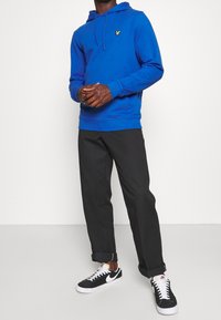 Persona che indossa una felpa blu con un logo giallo, pantaloni neri con risvolto e sneakers Nike bianco e nero, in piedi contro uno sfondo bianco semplice.