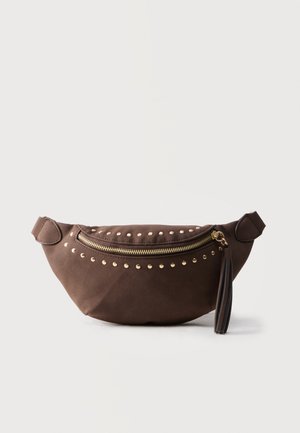 Sac ceinture en daim marron avec des clous dorés, une fermeture éclair dorée et un pompon, sur un fond uni clair.