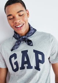 Camiseta de algodón gris con un logo de "GAP" en azul marino, combinada con una bandana oscura con patrón atada alrededor del cuello.