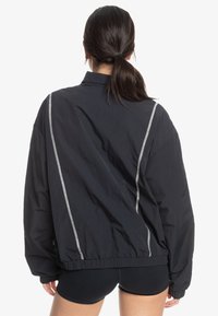 Svart lättviktsjacka med en lös passform, elastisk nederkant och kontrasterande vit sömnad längs ryggen och axlarna. Slät textur.