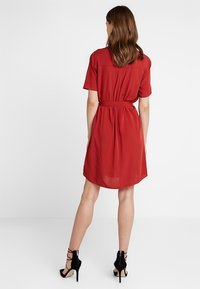 Vestito rosso a maniche corte con vita cinturata e design fluido. Presenta uno sprone posteriore e una texture leggermente trasparente. Abbinato a sandali neri con cinturini.