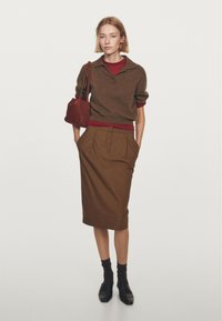 Kvinde står med hænderne i lommen iført brun nederdel, brun sweater over rød top, sorte sko og bærer en rød taske på skulderen.