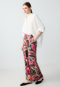 Camicia bianca ampia con colletto. Pantaloni a gamba larga colorati e decorati con grandi motivi a foglia in rosa, verde e giallo. Ballerine nere.