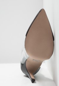 Chaussure à talon haut noire à bout pointu avec empeigne translucide et semelle beige texturée, photographiée en vue inférieure inclinée.