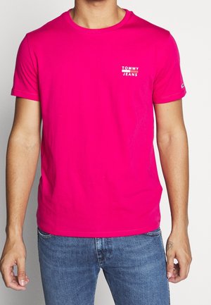 Homme portant un t-shirt Tommy Jeans rose vif à manches courtes et un jean bleu, debout devant un fond blanc uni.
