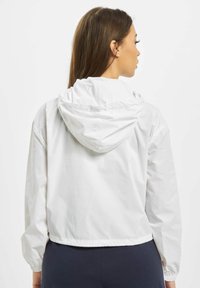 Valkoinen kevyt takki hupulla, lyhennetty malli, joustavat hihansuut ja sileä pinta. Ei näkyviä kuvioita tai metalliosia.