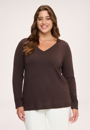 Femme souriante aux longs cheveux bruns, portant un pull marron foncé à col en V et un pantalon blanc, debout devant un fond clair.