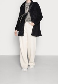 Schwarzer, texturierter Pelzmantel, weiße Hosen mit weitem Bein und kariertes Oberteil, kombiniert mit einer kleinen schwarzen Umhängetasche und weißen Turnschuhen.