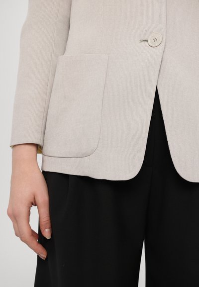 Beige ribbed jacket with a single button, featuring a front pocket and smooth texture, paired with black trousers.