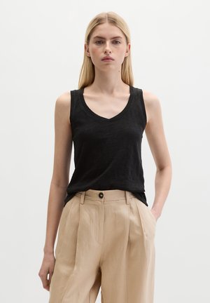 Frau mit glatten blonden Haaren trägt ein ärmelloses schwarzes V-Ausschnitt-Top und beige hoch taillierte Hosen, steht vor einem schlichten weißen Hintergrund.