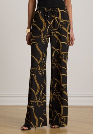 Zwarte wijdvallende broek met gouden ketting- en riemprint, gedragen met een zwarte top, sandalen en een gouden armband om de linker pols.