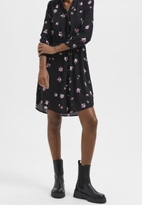 Vestido negro con estampado floral morado, de manga larga, con botones en la parte delantera y una ligera forma de A, combinado con botas negras hasta el tobillo.