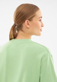 Jasnozielona bawełniana koszulka z okrągłym dekoltem i lekko opuszczonymi ramionami, gładka tekstura, widok z tyłu, włosy związane w kucyk.