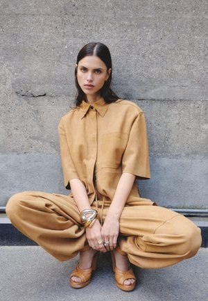 Tan suede jumpsuit with a buttoned collar and short sleeves; loose pants; paired with wooden platform sandals and layered bracelets.
