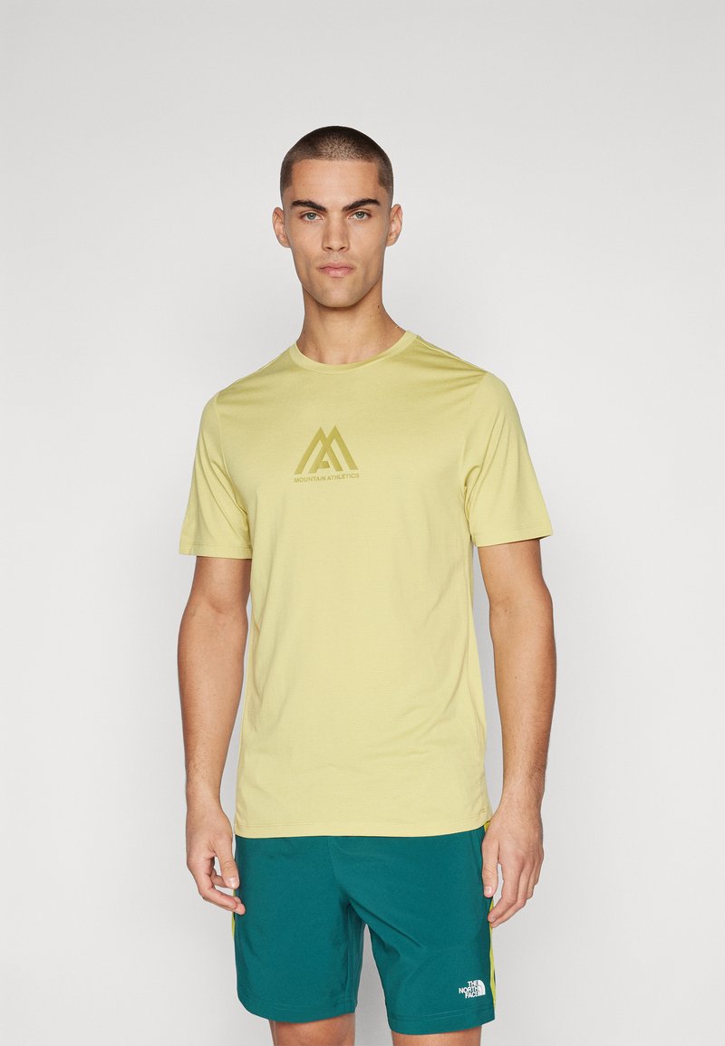 Trumpas rankovės šviesiai geltonas sportinis marškinėlis su apvaliu kaklu ir rudu Mountain Athletics logotipu krūtinėje. Lygi medžiagos tekstūra.