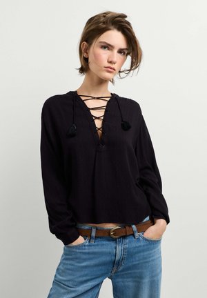 Jeune femme aux cheveux courts portant un chemisier noir à manches longues lacé, un jean bleu et une ceinture marron, debout les mains dans les poches devant un fond uni.