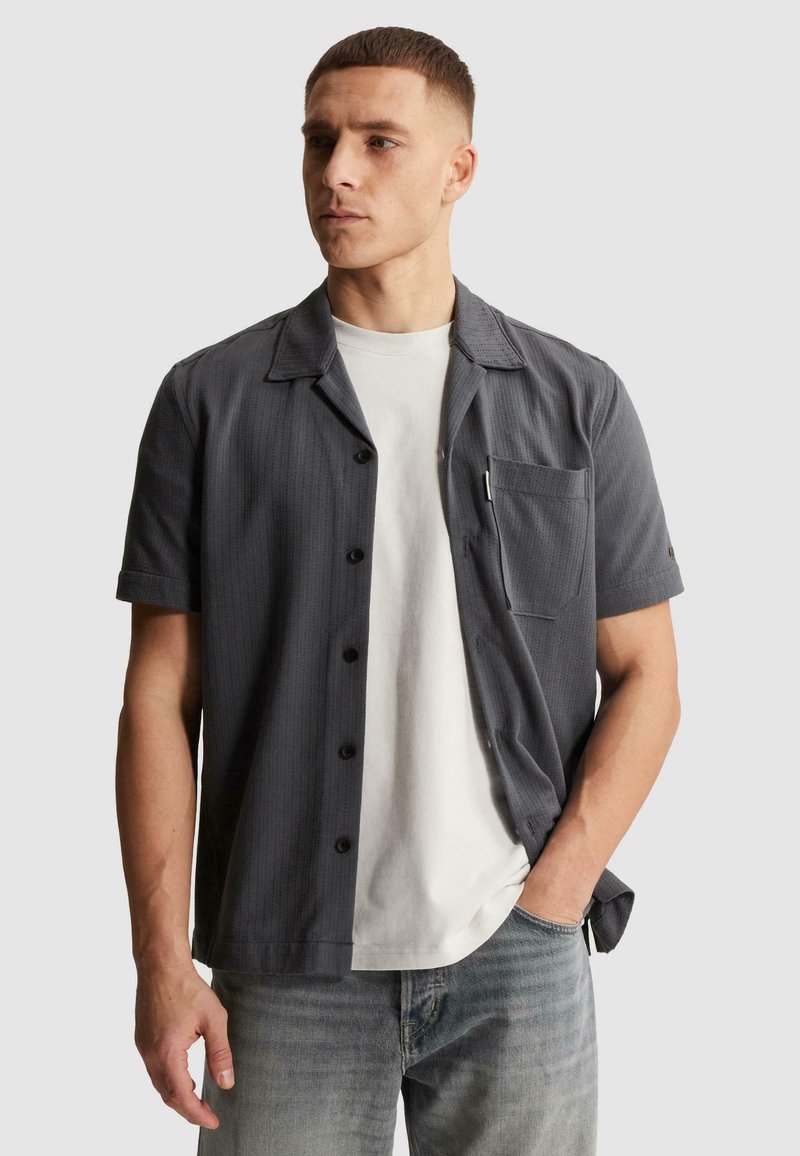 Junger Mann mit kurzen Haaren, der ein dunkelgraues kurzärmeliges Hemd über einem weißen T-Shirt und hellblauen Jeans trägt, Hand in der Tasche.