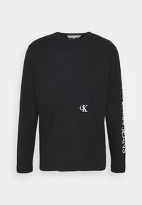 Černé dlouhé tričko s dlouhým rukávem vyrobené z bavlny. Obsahuje malé bílé logo "ck" na hrudi a text podél pravého rukávu.
