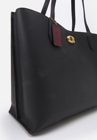 Black leather tote bag with a textured surface, gold hardware, and a maroon tag. Designed with two sturdy handles and a structured shape.