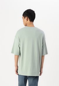 Lys grønn oversized T-skjorte med korte ermer, rund hals og myk bomullstekstur. Båret med blå denimjeans. Bakfra.