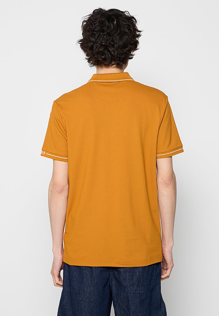 Persona con capelli ricci e scuri, che indossa una polo gialla senape a maniche corte e pantaloni in denim blu scuro, di spalle.