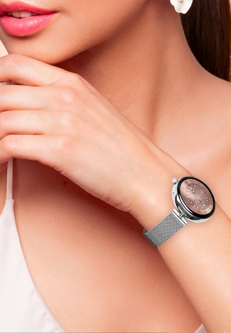 Montre ronde avec un bracelet en maille argentée, dotée d'un cadran en or rose avec un design découpé complexe. Main positionnée près du cadran.