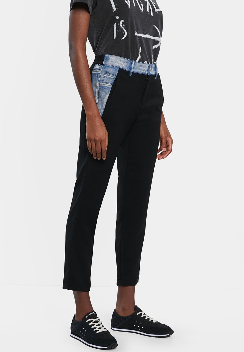 Personne portant un pantalon noir avec un détail de ceinture en denim bleu, un t-shirt noir imprimé et des baskets noires avec des lacets blancs.