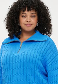 Pulover tricotat de culoare albastră, cu un design texturat de sârmă, având un guler cu fermoar pe jumătate și mâneci largi. Accesoriile fermoarului sunt de culoare argintie.