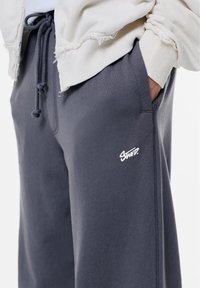 Grijze katoenen sweatpants met een zachte textuur, een tailleband met trekkoord, zijzakken en een wit logo op de linkerdij. Gecropte weergave met een licht jack.