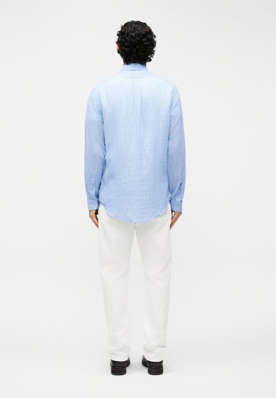 Άνδρας με σγουρά σκούρα μαλλιά στέκεται με την πλάτη, φοράει ανοιχτό μπλε ριγέ πουκάμισο μακρυμάνικο, άσπρο παντελόνι και σκούρα παπούτσια σε απλό φόντο.