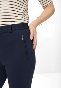 Marineblaue dehnbare Hosen verfügen über einen strukturierten Bund und eine Reißverschlusstasche an der Seite, mit einem insgesamt glatten, schlichten Stoff.