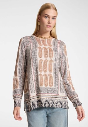 Vrouw draagt een blouse met lange mouwen en paisley-patroon in beige, bruin en zwart, met lichtblauwe spijkerbroek tegen een effen achtergrond.