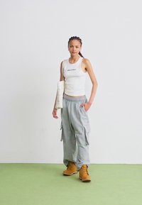 Hvid tanktop med teksten "Dickies", grå cargo bukser og gule støvler. Modellen holder en hvid tekstureret taske. Enkel baggrund og grøn gulv.