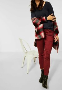 Femme portant un haut noir, une veste à carreaux rouges, un pantalon bordeaux et des bottines noires, debout à côté d'une chaise métallique blanche sur un fond uni.