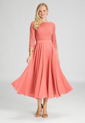 Robe couleur corail rose avec des manches longues en dentelle, corsage ajusté et jupe fluide. Ourlet évasé, drapé élégant, complété par des accents dorés sur les sandales.