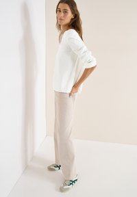 Femme aux longs cheveux bruns vue de profil, portant un pull blanc texturé, un pantalon beige et des baskets grises avec des rayures vertes.