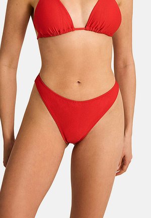 Femme portant un haut de bikini triangle rouge texturé et un bas de bikini taille haute assorti sur un fond clair uni.