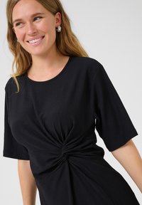 Robe noire à manches courtes, en tissu texturé, avec un détail noué à la taille. Encolure ronde simple ; matière douce et légère.
