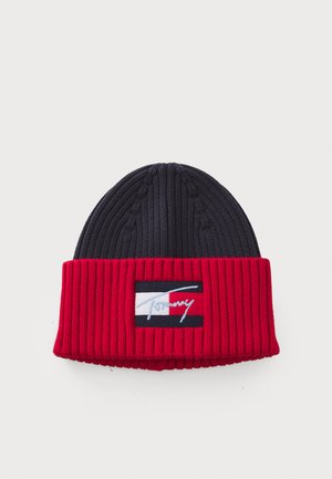 Bonnet en tricot bleu marine et rouge, avec une texture côtelée et un revers replié. Comprend une étiquette rectangulaire avec le logo de la marque en bleu clair.