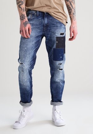 Homme portant un jean bleu déchiré avec des patchs foncés, une chemise beige, des baskets blanches et des bras tatoués sur un fond uni.