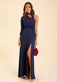 Robe maxi bleu marine avec une longue manche, décolleté drapé, fente haute sur le côté et un détail noué à l'épaule. Talons compensés dorés et une pochette rouge.