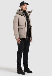 Beige puffer jacket with a hood, front pockets, and a zipper closure, paired with black jeans and black lace-up boots.