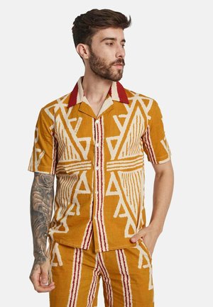 Chemise à manches courtes de couleur jaune doré avec des motifs tribaux blancs et rouges, dotée d'un col et de boutons rouges contrastants. Fabriquée en tissu léger.