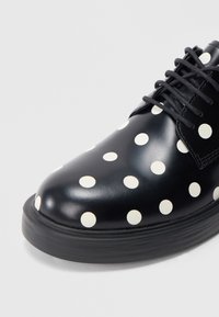 Sort lædersko med hvide polkaprikker, rund tå, glat tekstur og sorte snørebånd; med en tyk, tekstureret sål.
