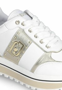 Sneaker bianca con accenti metallici argento, caratterizzata da un'etichetta con il marchio, borchie decorative e una suola in gomma spessa con trazione.