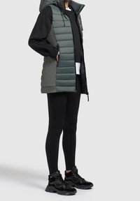 Piumino verde imbottito con collo alto, indossato sopra una felpa nera e leggings neri, completato da sneakers nere con dettagli testurizzati.